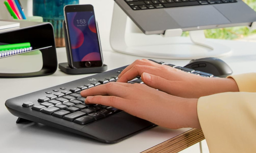 Osoba pisze na grafitowej klawiaturze z logo \'Logitech\', obok znajduje się myszka i stojący telefon z wyświetlaczem pokazującym godzinę 1:53.