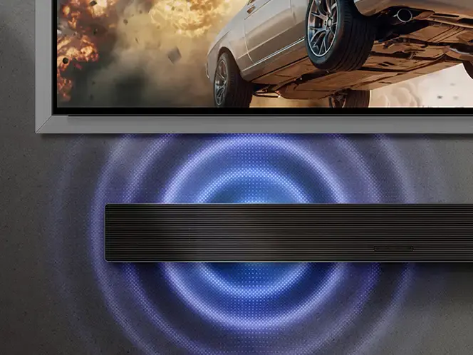 2026-hw-qs90h-ai-dynamic-bass-control 664x498 Na obrazie widoczny jest soundbar umieszczony pod telewizorem, z którego wydobywają się niebieskie fale dźwiękowe. Na ekranie telewizora widać fragment samochodu w dynamicznej scenie z eksplozją.