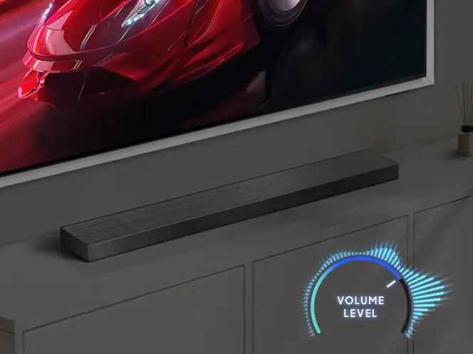 Na obrazie widoczny jest soundbar w kolorze grafitowo-czarnym umieszczony na szafce pod telewizorem. Obok soundbara znajduje się graficzny wskaźnik z napisem \'VOLUME LEVEL\'.