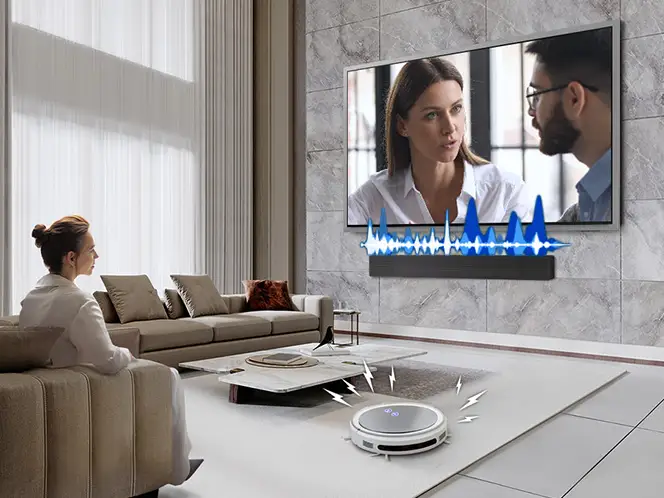 W nowoczesnym salonie znajduje się duży telewizor z obrazem przedstawiającym dwie osoby oraz soundbar zamontowany pod nim. Na podłodze widoczny jest okrągły robot sprzątający.
