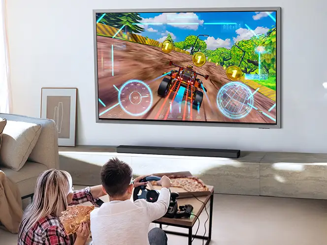 2026-hw-qs90h-game-pro-mode 664x498 Dwie osoby siedzą na kanapie przed dużym telewizorem, na którym wyświetlana jest gra wyścigowa. Przed telewizorem znajduje się soundbar, a na stoliku leży kontroler do gier i kawałki pizzy.