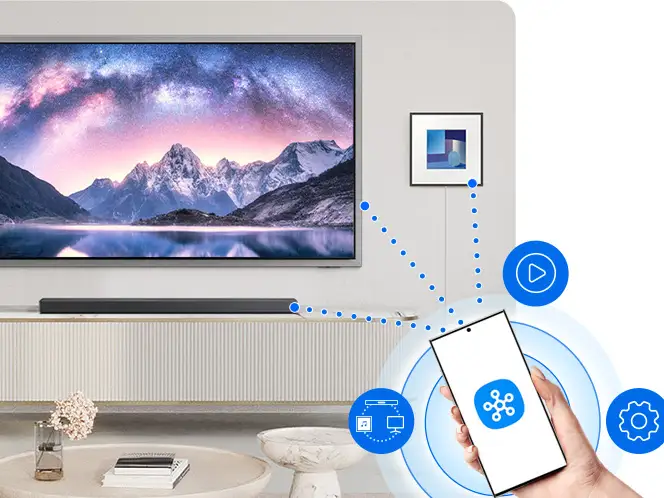 Na obrazie widać nowoczesny salon z dużym telewizorem wyświetlającym krajobraz górski. Przed telewizorem znajduje się soundbar, a obok ręka trzymająca smartfon z ikoną aplikacji, z którego wychodzą linie wskazujące na różne funkcje multimedialne.