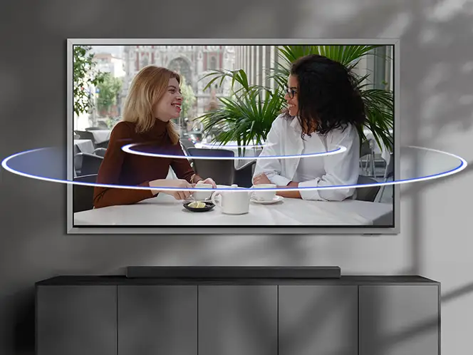 Na obrazie widoczny jest telewizor z wyświetlanym obrazem dwóch osób siedzących przy stole w jasnym pomieszczeniu z roślinami w tle. Przed telewizorem znajduje się soundbar, a wokół niego widoczne są efekty świetlne sugerujące dźwięk przestrzenny.