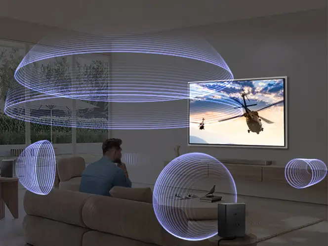 Osoba siedzi na kanapie w salonie, oglądając telewizor z wyświetlanym obrazem helikoptera na tle nieba. Wokół rozchodzą się wizualizacje fal dźwiękowych, sugerujące działanie systemu dźwiękowego.