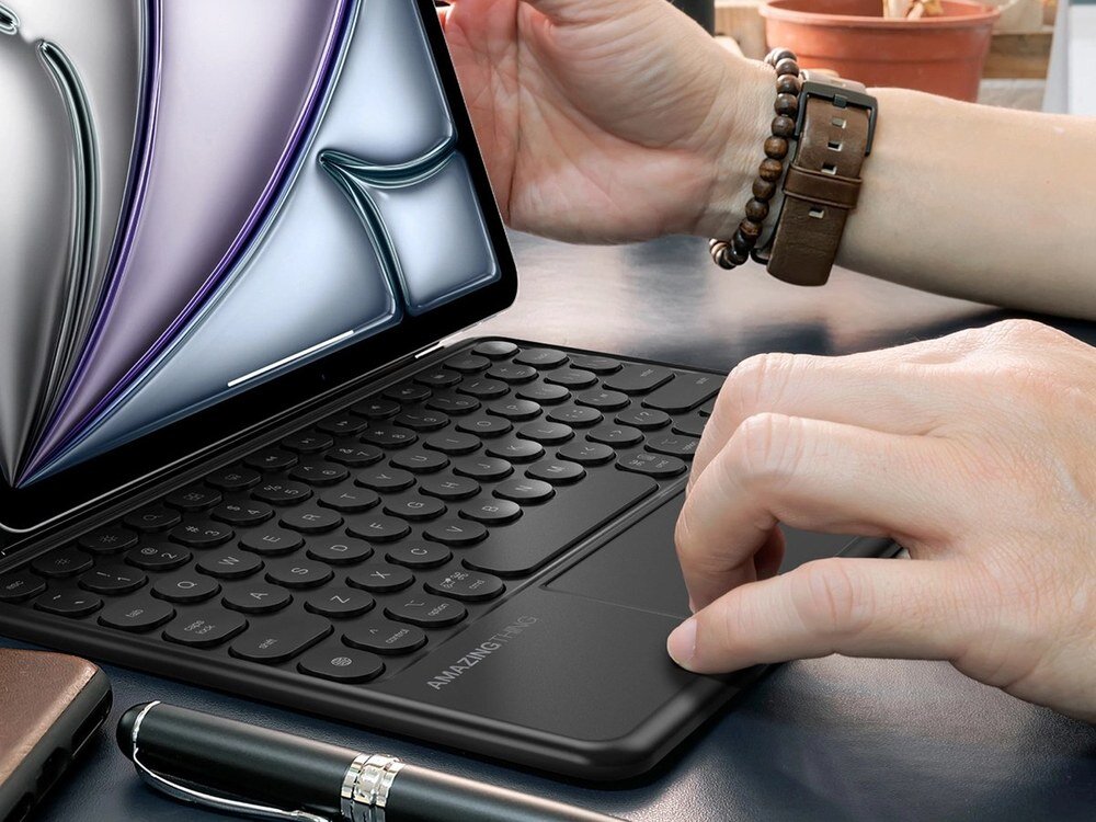Zbliżenie na czarną klawiaturę AmazingThing z okrągłymi klawiszami i touchpadem — dłonie użytkownika obsługują touchpad w pobliżu iPada Air.