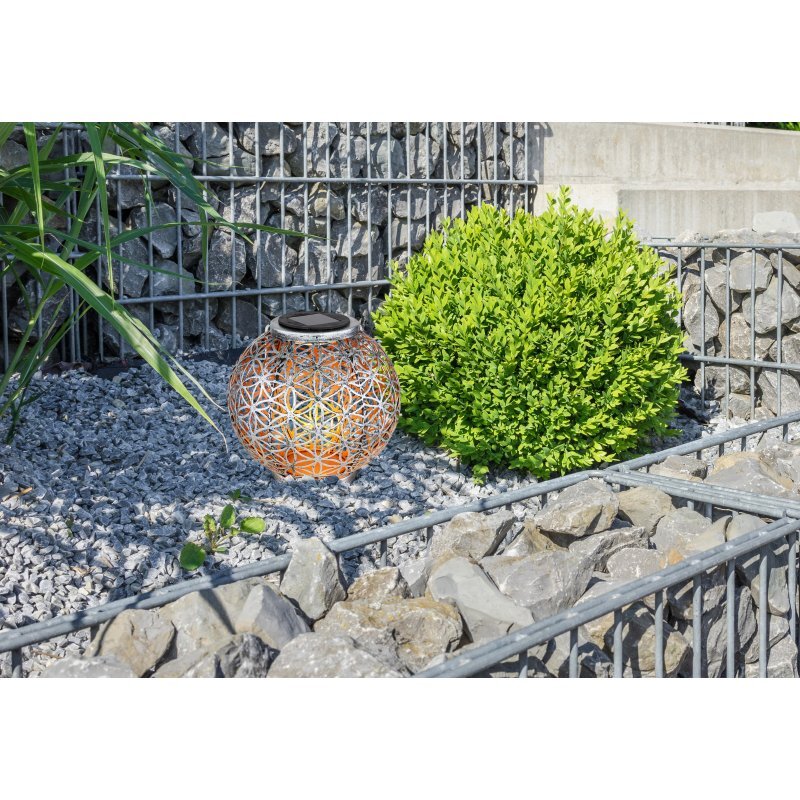 Na obrazie widoczna jest ozdobna lampa solarna o ażurowym wzorze, umieszczona na żwirowym podłożu obok zielonego krzewu. W tle znajdują się kamienie i metalowa siatka.