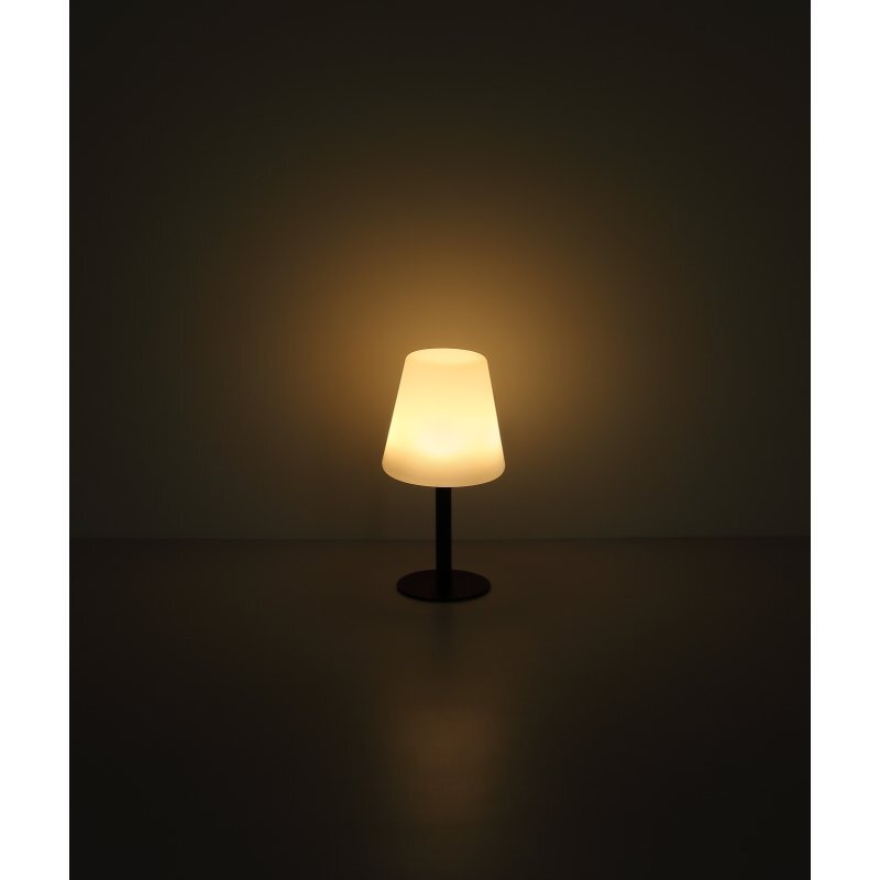 Na obrazie widoczna jest lampa stołowa z białym kloszem, emitująca ciepłe, żółte światło. Lampa stoi na ciemnej podstawie, a tło jest ciemne.