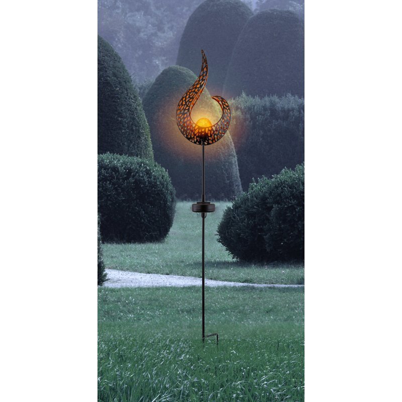 Lampa solarna LED w kształcie płomienia, umieszczona na metalowym pręcie, oświetla otoczenie ciepłym światłem. W tle widoczne są przycięte krzewy i trawnik.