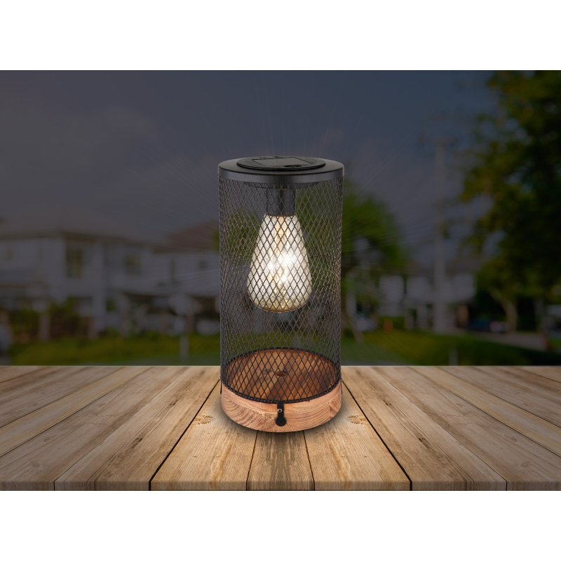 Na drewnianym stole stoi lampa solarna w kształcie walca z siatkowym kloszem, wewnątrz której znajduje się świecąca żarówka. W tle widoczny jest rozmyty obraz ogrodu i budynków.