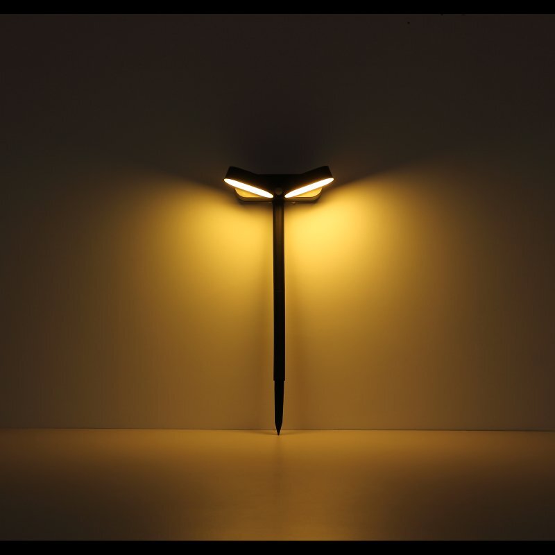 Lampa solarna LED emituje ciepłe, żółte światło, tworząc symetryczne rozświetlenie na ścianie. Lampa jest zamontowana na ciemnym, smukłym trzonku.
