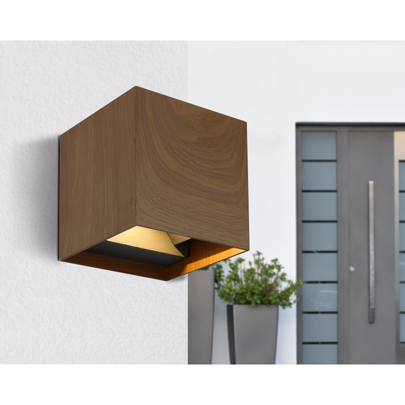 Na obrazie widoczna jest lampa ścienna o kwadratowym kształcie, imitująca drewno, zamontowana na białej ścianie. W tle znajduje się nowoczesne wejście do budynku z dużymi donicami z roślinami.