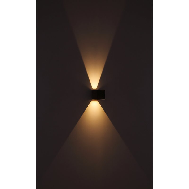Na obrazie widoczna jest lampa ścienna emitująca ciepłe światło skierowane w górę i w dół, tworząc efektowne promienie na ścianie. Lampa ma prostokątny kształt i ciemny kolor, przypominający drewno.