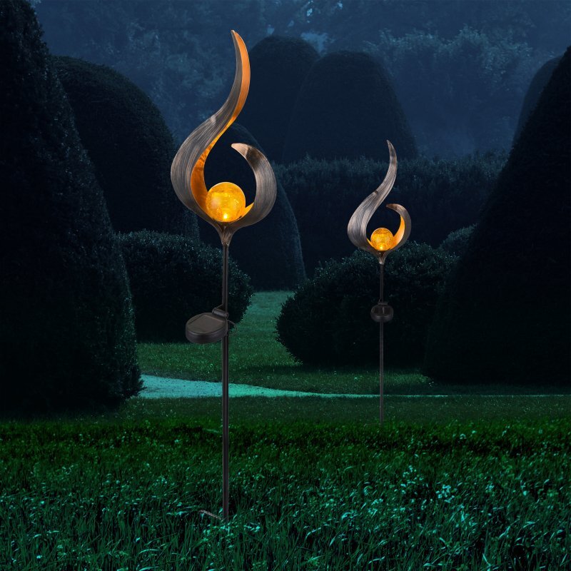 Na obrazie widoczne są dwie lampy solarne o dekoracyjnym kształcie, emitujące ciepłe, żółte światło. Lampy umieszczone są na tle ciemnego ogrodu z przyciętymi krzewami.