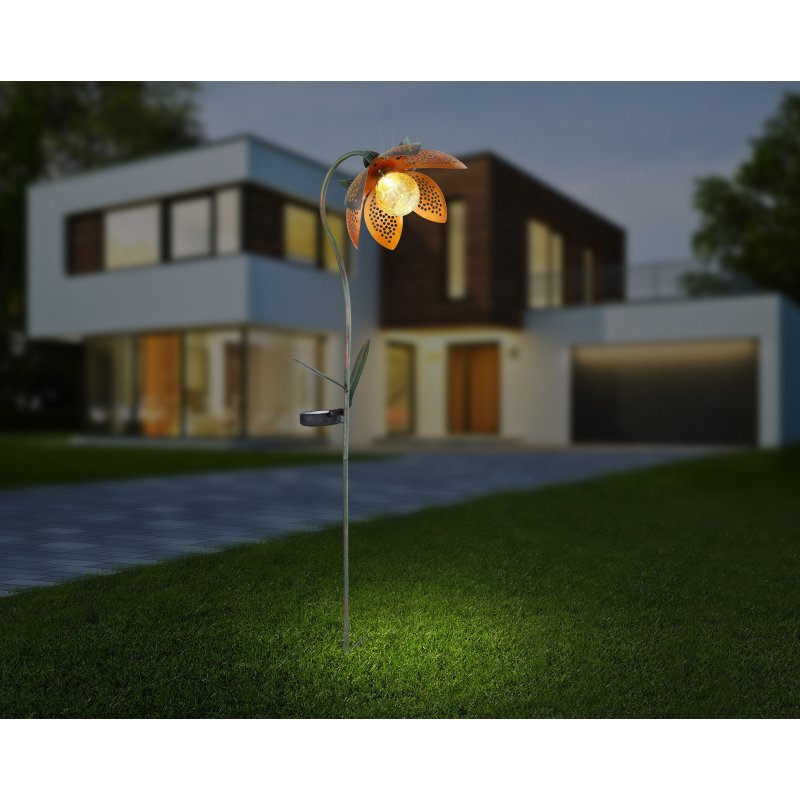 Na trawniku przed nowoczesnym domem znajduje się lampa solarna w kształcie kwiatu z pomarańczowymi płatkami i świecącym środkiem. W tle widać oświetlony budynek z dużymi oknami.