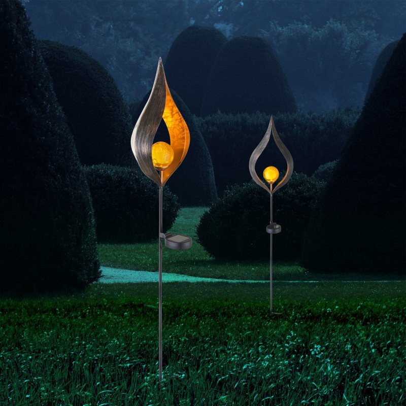 Na obrazie widoczne są dwie lampy solarne o kształcie przypominającym płomień, z żółtym światłem w środku. Lampy umieszczone są na tle ogrodu z przyciętymi krzewami.