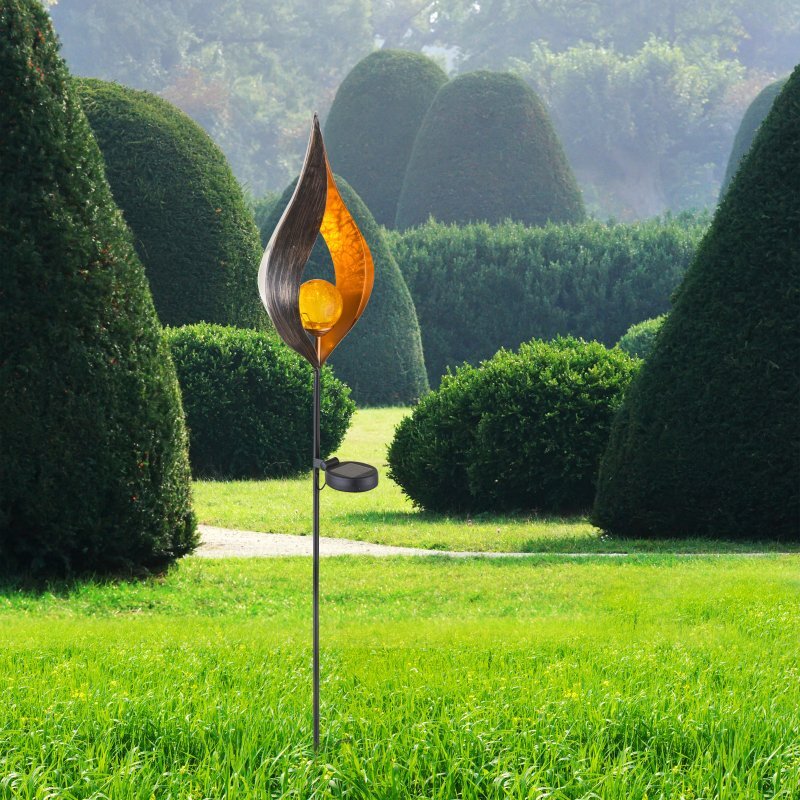 Na obrazie widoczna jest lampa solarna z dekoracyjnym kloszem w kształcie płomienia, umieszczona na tle ogrodu z przyciętymi krzewami. Lampa ma metaliczny wygląd z pomarańczowym wnętrzem i kulistym źródłem światła.