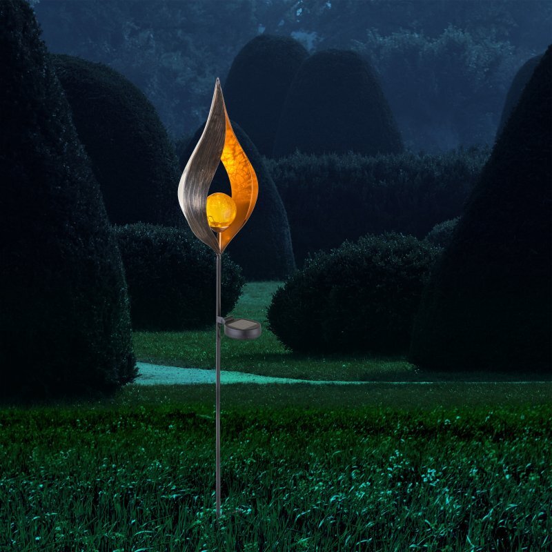 Lampa solarna o kształcie przypominającym płomień, z żółtym światłem wewnątrz, umieszczona na tle ogrodu z przyciętymi krzewami.