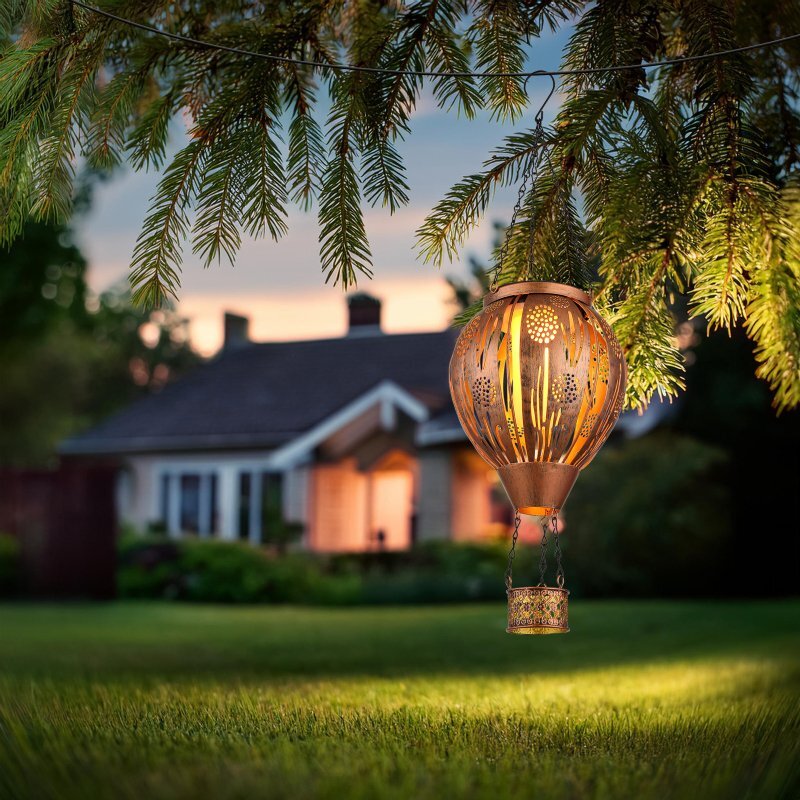 Na obrazie widać dekoracyjną lampę solarną w kształcie balonu na ogrzane powietrze, wiszącą na tle ogrodu z domem w oddali. Lampa emituje ciepłe światło, a wokół niej znajdują się gałęzie drzewa.
