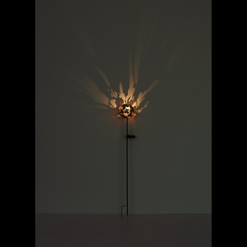Lampa solarna LED zamontowana na długim pręcie, rzucająca dekoracyjne cienie na ścianę. Światło tworzy wzory przypominające liście lub gałęzie.