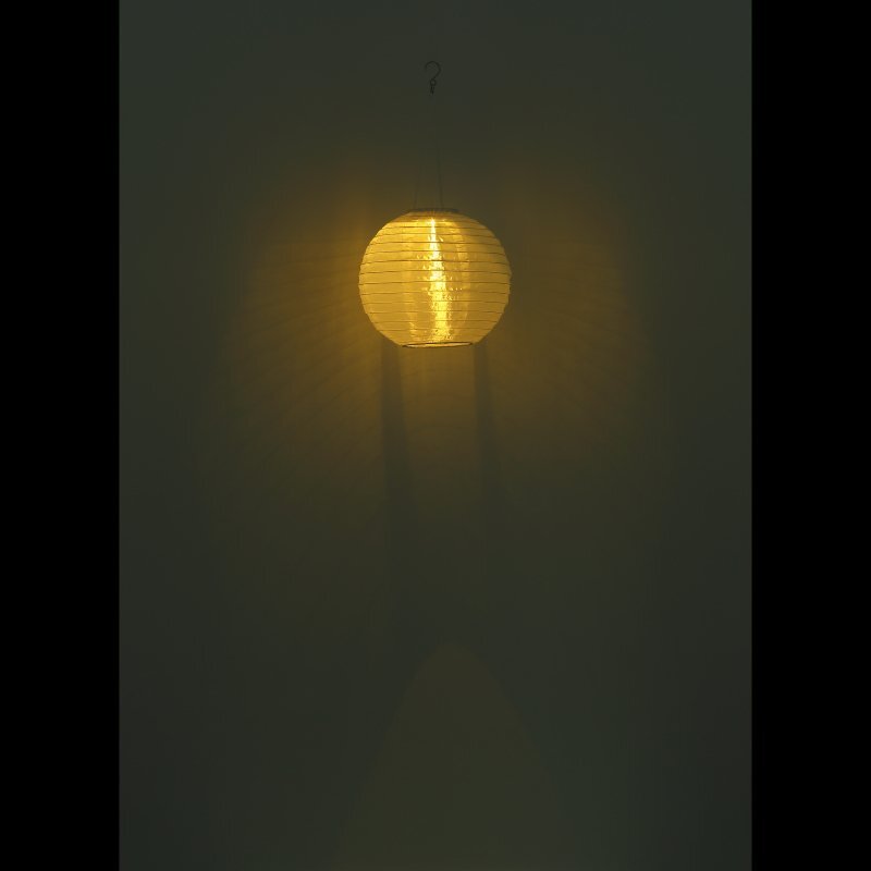 Na ciemnym tle wisi okrągła lampa emitująca ciepłe, żółte światło. Lampa ma strukturę przypominającą papierowy lampion.