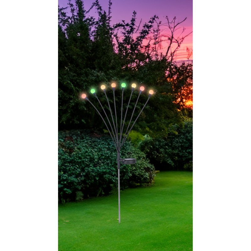 Na obrazie widoczna jest lampa solarna ogrodowa z ośmioma kolorowymi diodami LED, umieszczona na trawniku. W tle znajduje się zieleń krzewów i drzewa na tle kolorowego nieba o zachodzie słońca.