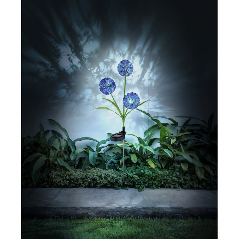 Na obrazie widoczna jest lampa solarna LED w kształcie trzech niebieskich kwiatów, umieszczona w ogrodzie wśród zielonych roślin. Lampa emituje światło, tworząc wzory na ścianie w tle.