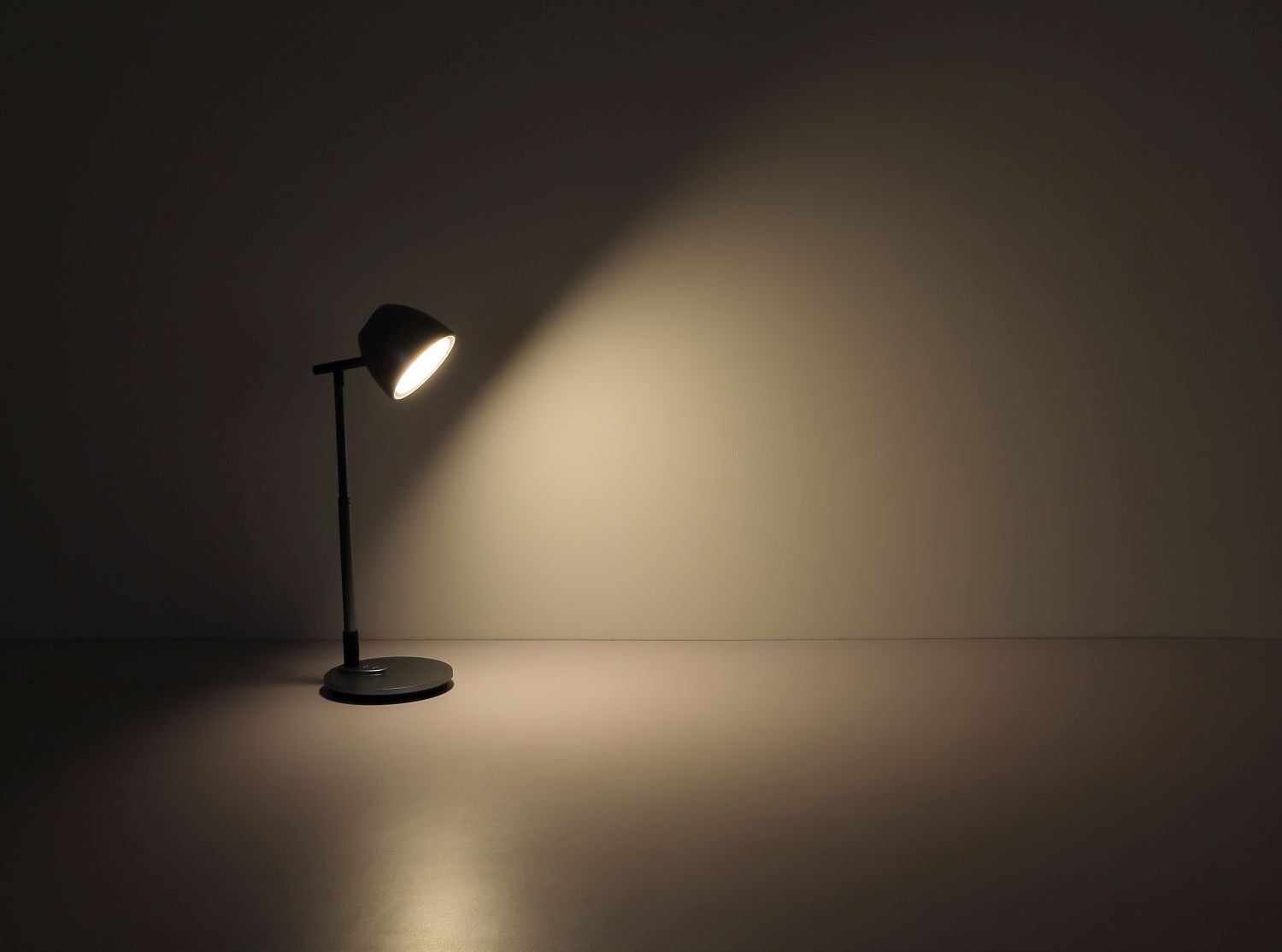 Na obrazie widoczna jest czarna lampa biurkowa, która oświetla ścianę i powierzchnię stołu ciepłym światłem. Lampa ma prosty, nowoczesny design z okrągłą podstawą i regulowanym ramieniem.
