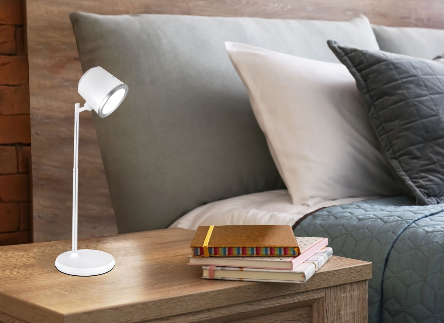 Na drewnianym stoliku nocnym stoi biała lampa biurkowa LED o nowoczesnym designie. Obok lampy leży stos kolorowych książek, a w tle widoczna jest część łóżka z poduszkami i narzutą.