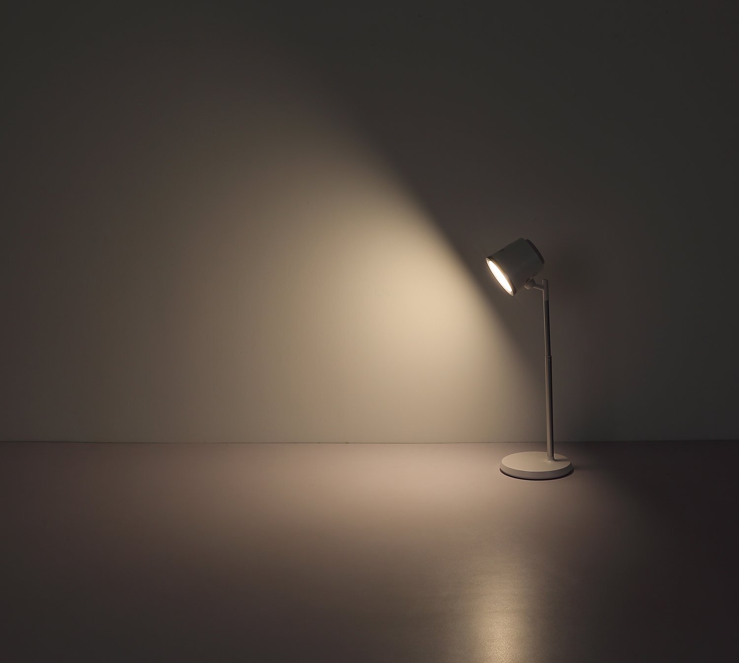 Biała lampa biurkowa LED stoi na płaskiej powierzchni, emitując światło na ścianę, tworząc delikatny, rozproszony blask.