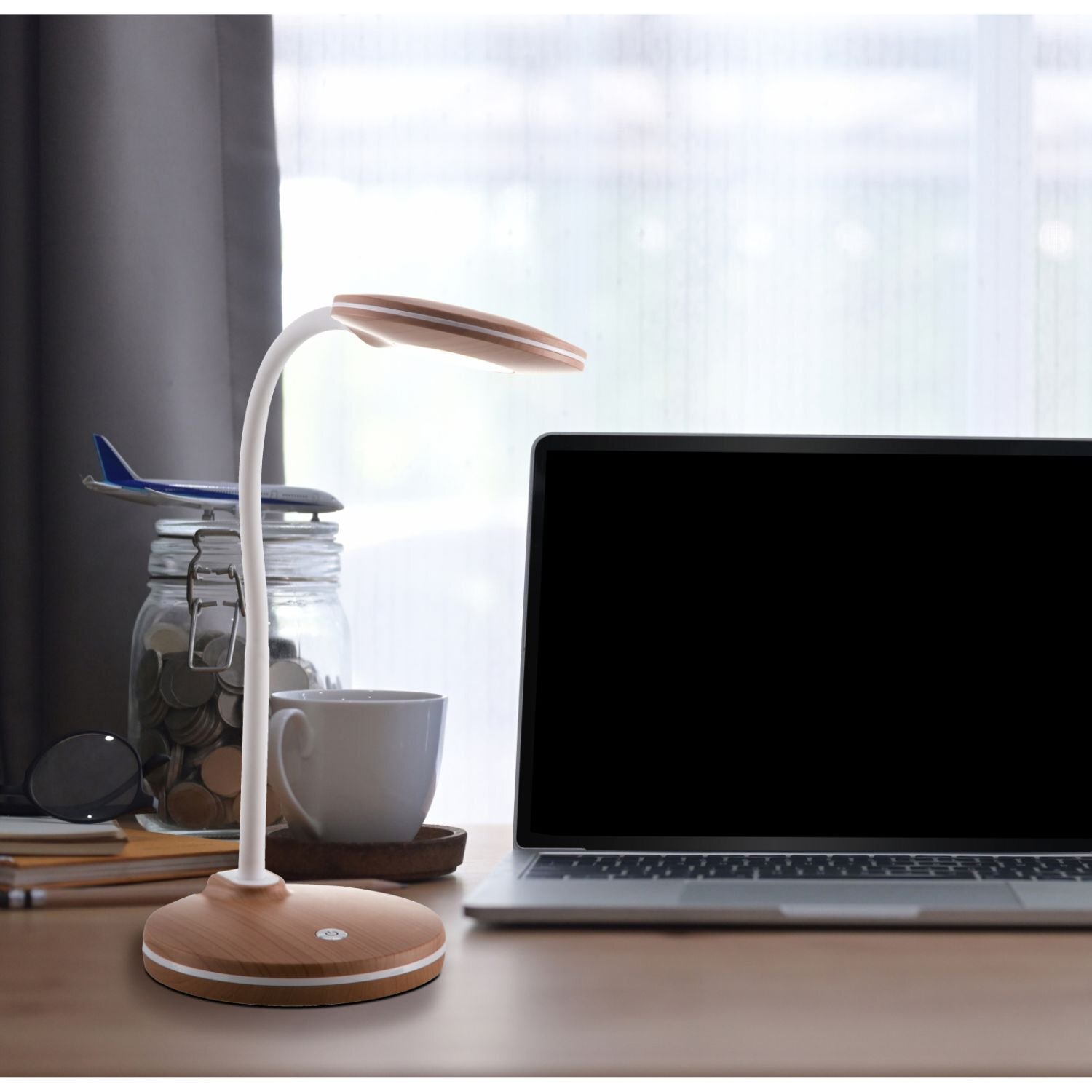 Na biurku znajduje się nowoczesna lampa biurkowa z drewnianą podstawą i białym, elastycznym ramieniem. Obok lampy stoi laptop, filiżanka oraz słoik z monetami i model samolotu.