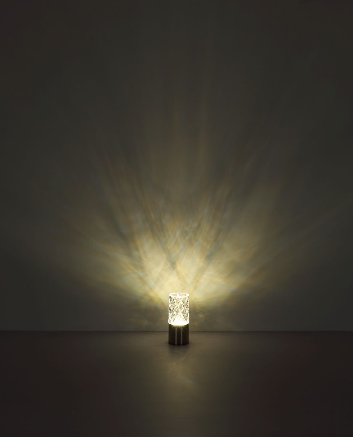 Na ciemnym tle znajduje się włączona lampa stołowa, emitująca rozproszone światło w kształcie promieni. Lampa ma cylindryczny kształt z dekoracyjnym wzorem na powierzchni.