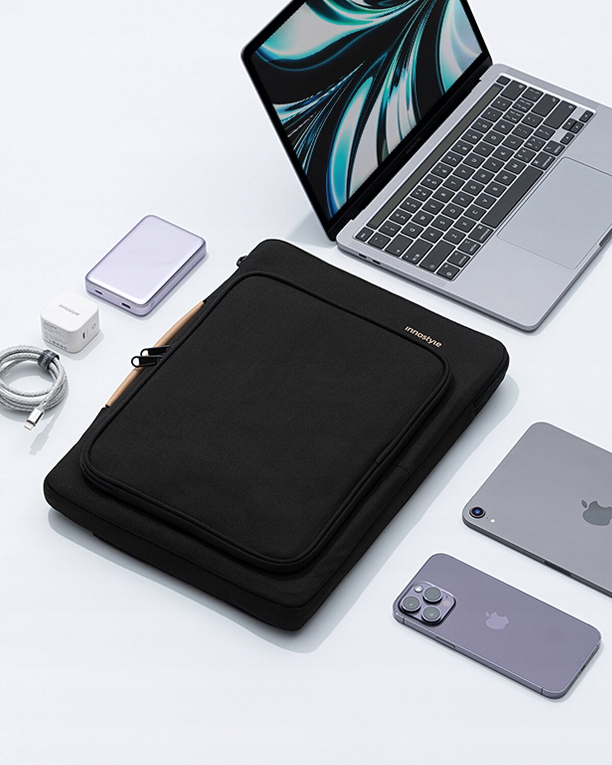 Na obrazie widoczna jest czarna torba na laptopa z napisem \'innostyle\', otoczona różnymi urządzeniami elektronicznymi, w tym laptopem, smartfonem i tabletem. Torba ma dodatkową kieszeń z przodu i jest umieszczona na białym tle.
