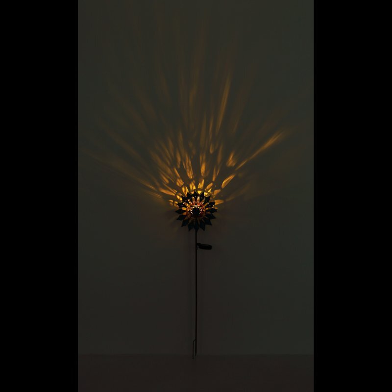 Lampa solarna LED emituje ciepłe, rozproszone światło, tworząc wzór przypominający promienie słońca na ścianie. Lampa jest zamontowana na długim pręcie, co sugeruje jej przeznaczenie do ogrodu.