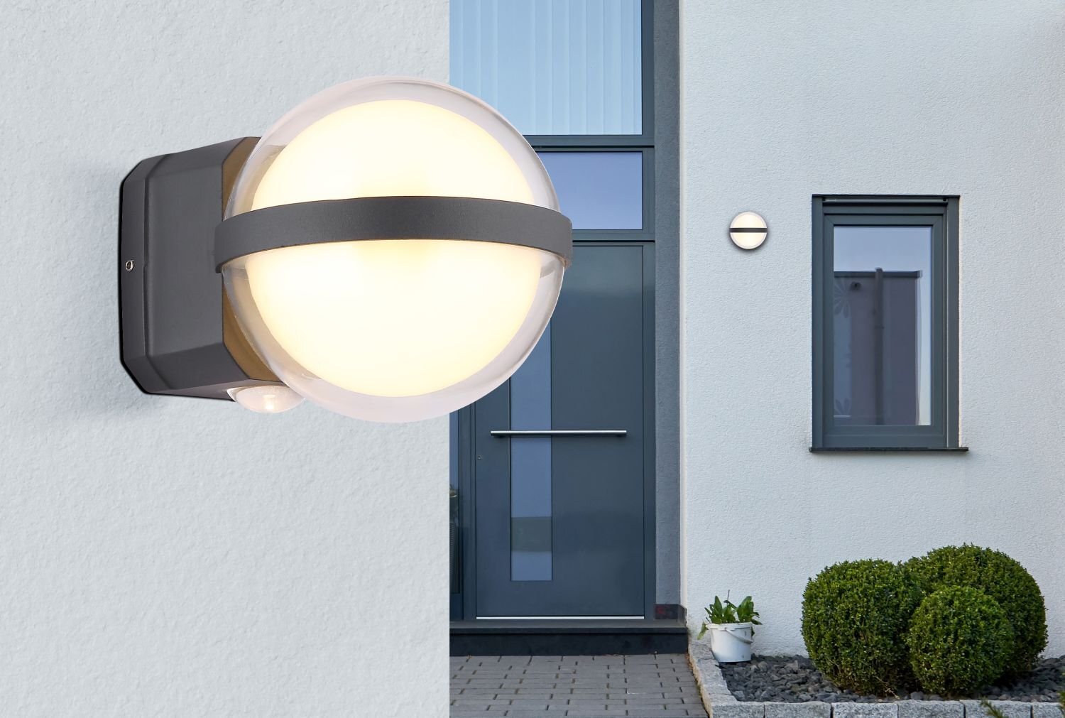 Na obrazie widoczna jest nowoczesna lampa ścienna LED o kulistym kształcie, zamontowana na zewnętrznej ścianie budynku. W tle znajduje się wejście do domu z dużymi drzwiami i oknem, a obok rosną krzewy w donicach.