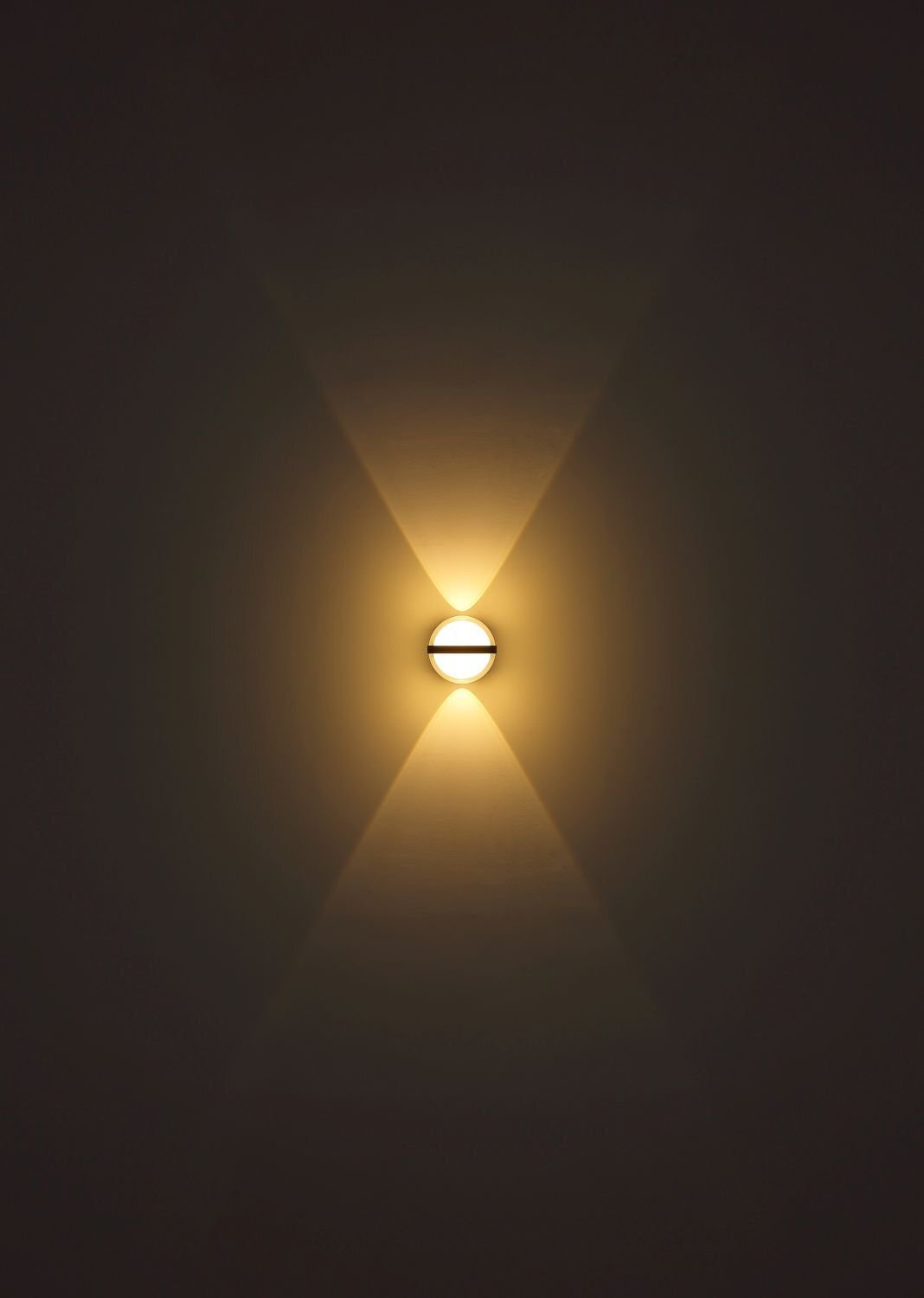 Na obrazie widoczna jest lampa ścienna emitująca ciepłe, rozproszone światło w kształcie dwóch stożków skierowanych w górę i w dół. Światło tworzy symetryczny wzór na ciemnej ścianie.