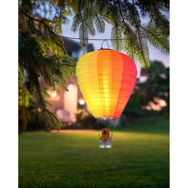 Na obrazie widoczna jest wisząca lampa solarna w kształcie balonu, świecąca ciepłym, żółto-pomarańczowym światłem. W tle znajduje się ogród z drzewami i domem.