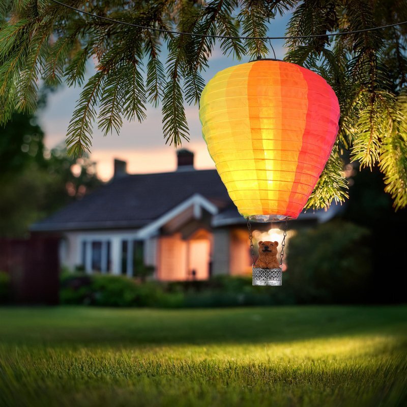 Lampa solarna w kształcie balonu na gorące powietrze wisi na tle ogrodu z domem. Balon jest kolorowy, a w koszu znajduje się figurka misia.