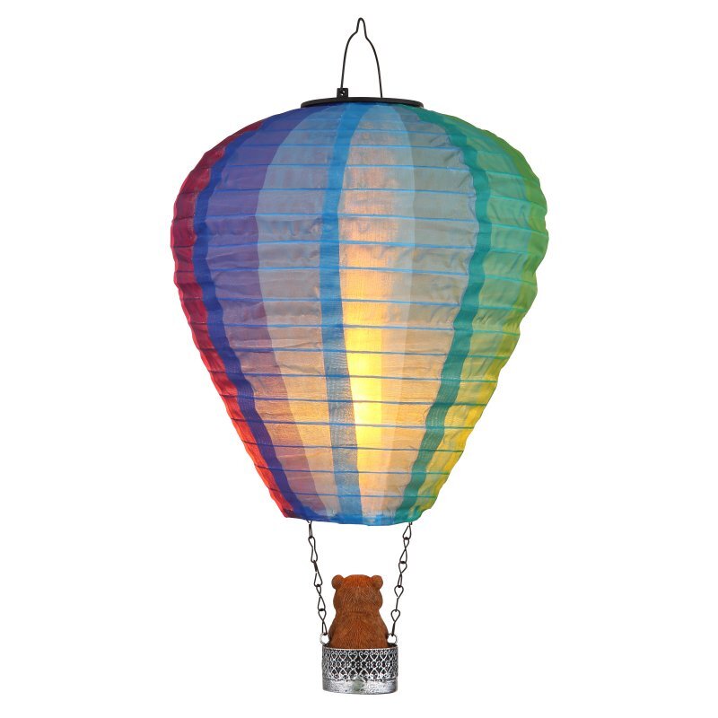 Kolorowa lampa solarna w kształcie balonu z koszem, w którym znajduje się figurka misia. Lampa emituje ciepłe światło, a jej powierzchnia jest pokryta pionowymi pasami w różnych kolorach tęczy.