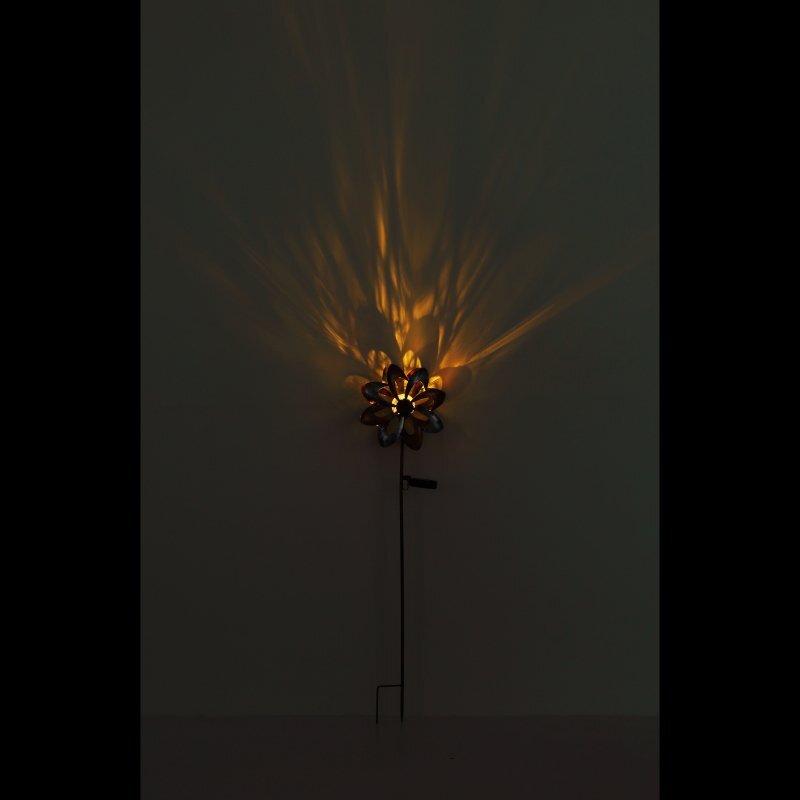 Na ciemnym tle widoczna jest lampa solarna w kształcie kwiatu, emitująca ciepłe, żółte światło, które tworzy wzory na ścianie.