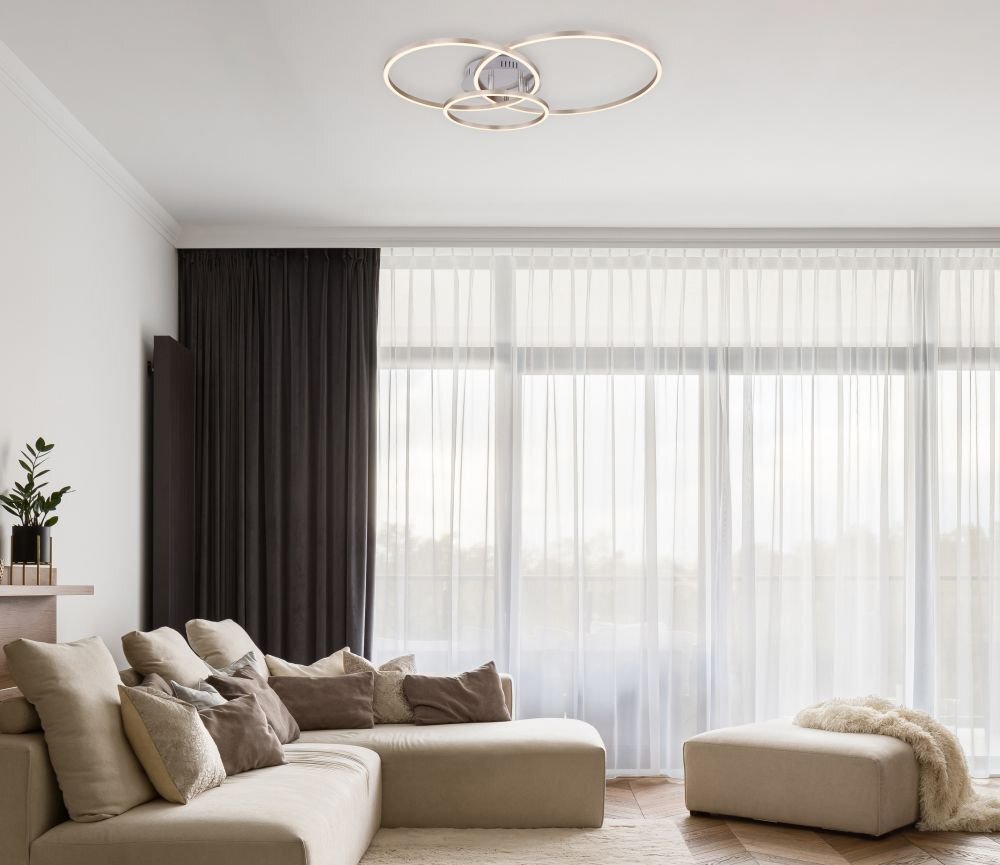 W nowoczesnym salonie znajduje się lampa sufitowa z trzema okrągłymi pierścieniami. Wnętrze jest jasne, z dużymi oknami i zasłonami, a na sofie leżą poduszki.