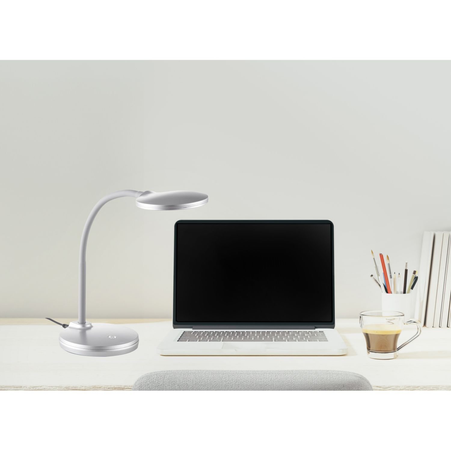 Na biurku znajduje się srebrna lampa biurkowa LED obok otwartego laptopa z czarnym ekranem. Obok laptopa stoi szklanka z kawą oraz pojemnik z przyborami do pisania.
