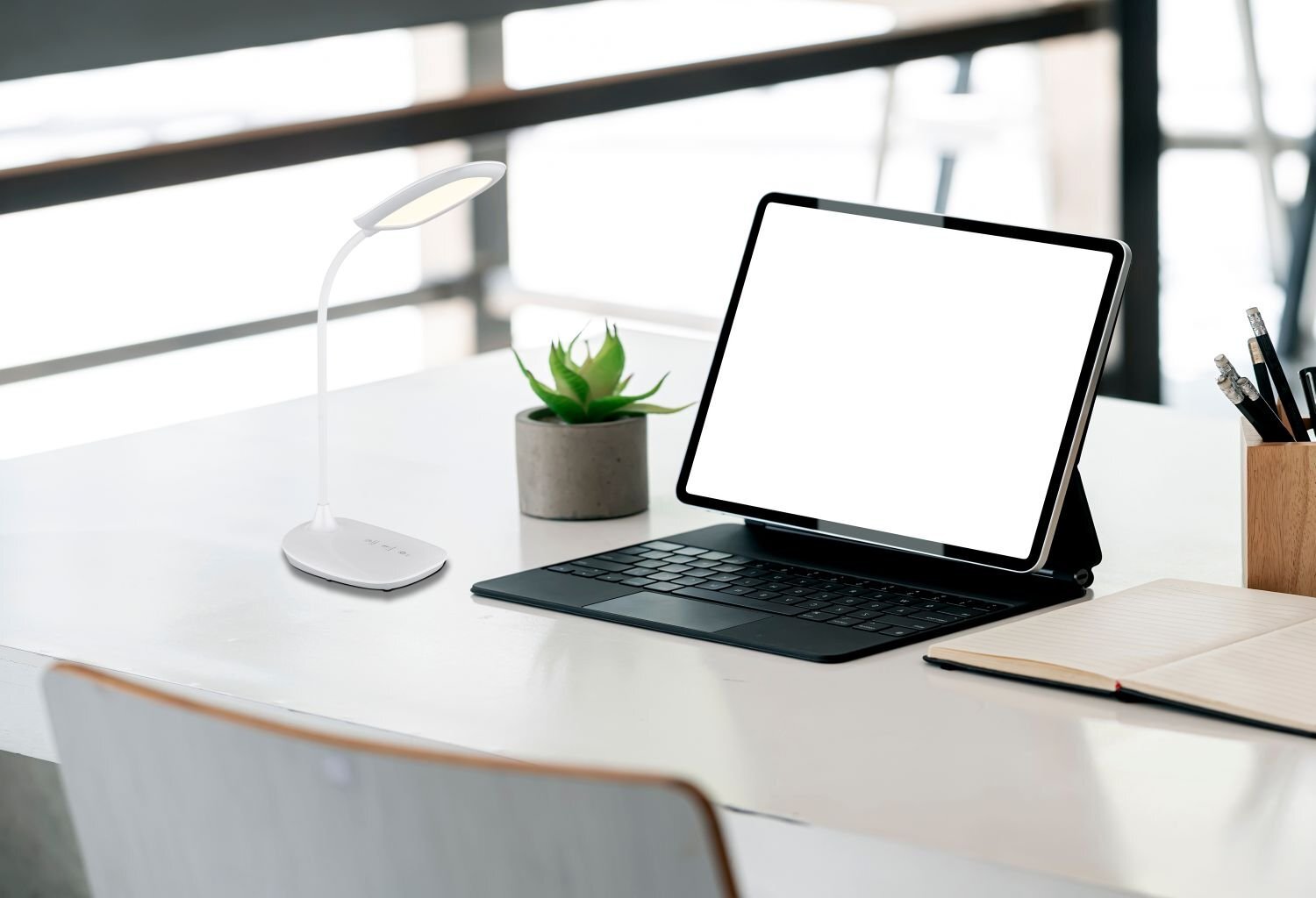 Na biurku znajduje się nowoczesna biała lampka biurkowa LED oraz tablet z klawiaturą. Obok lampki stoi mała roślina w doniczce, a na biurku leży otwarty notes z długopisem.