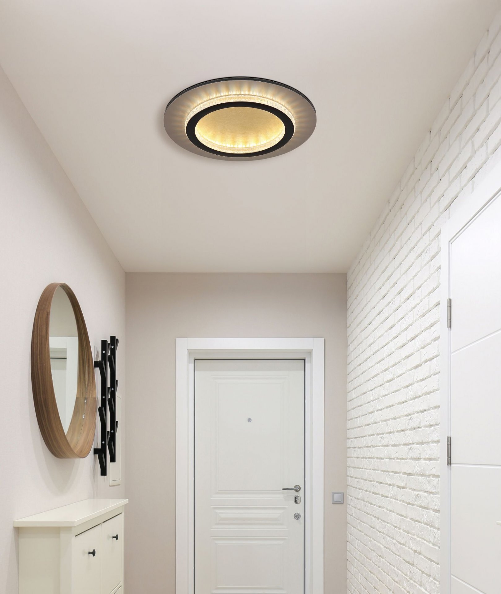 Na suficie znajduje się nowoczesna lampa sufitowa LED o okrągłym kształcie, emitująca ciepłe światło. Wnętrze pomieszczenia jest jasne, z białymi drzwiami i ścianą z białej cegły.