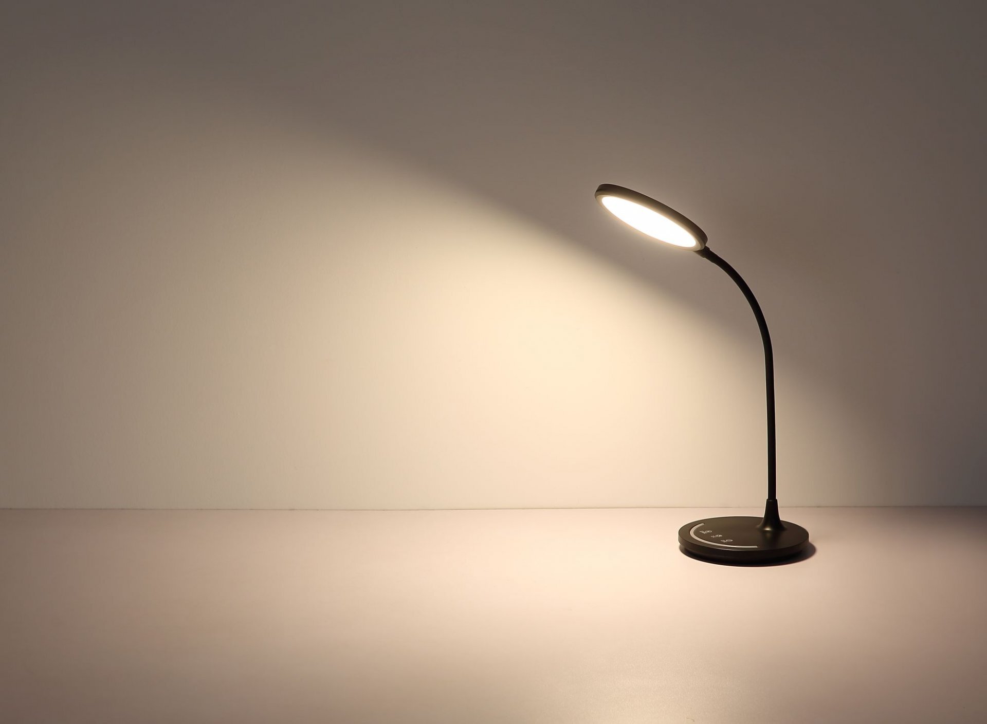 Na obrazie widoczna jest nowoczesna lampa biurkowa LED o czarnym kolorze, emitująca ciepłe światło na jasne tło. Lampa ma elastyczne ramię i okrągłą podstawę.
