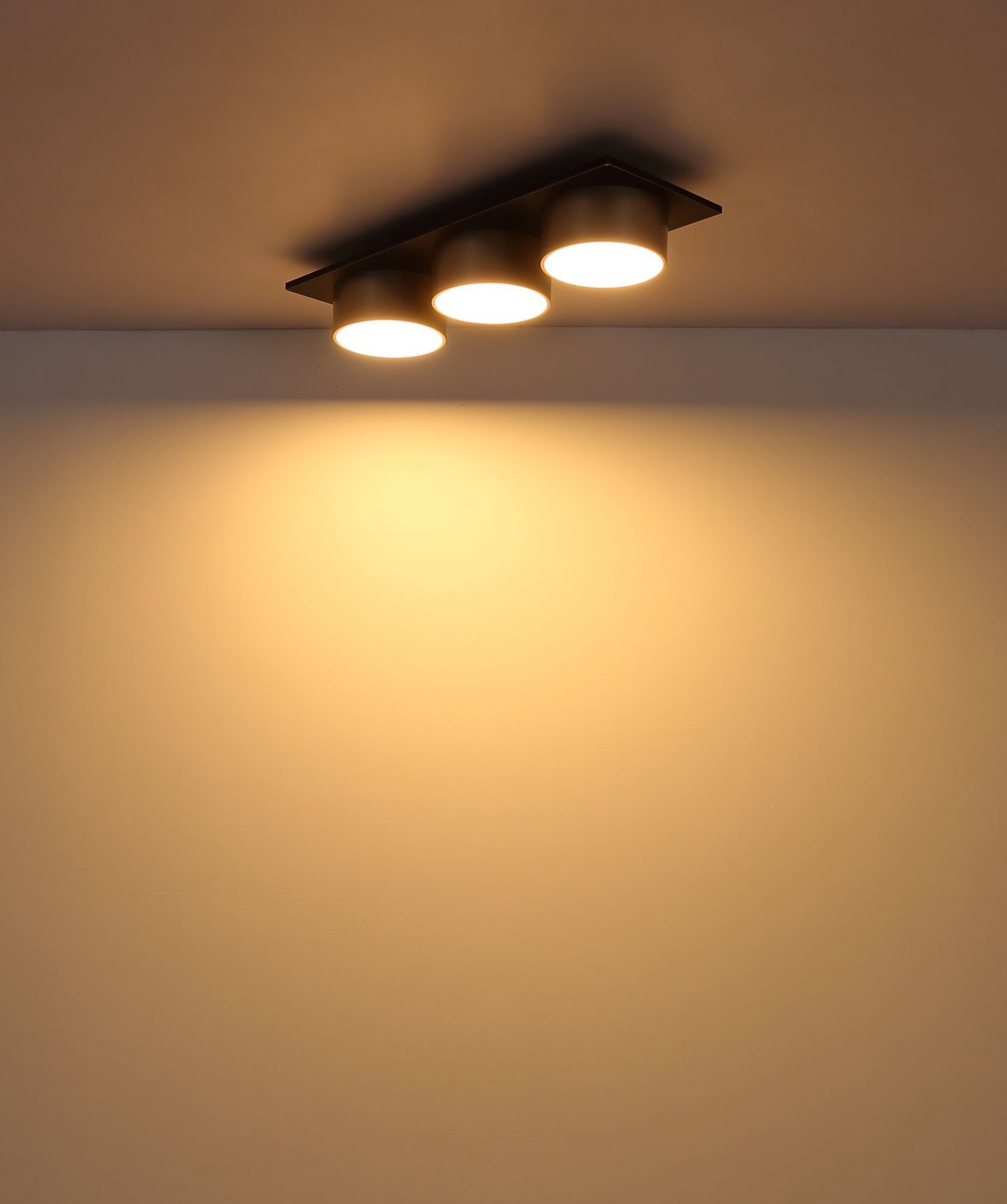 Na suficie zamontowana jest nowoczesna lampa sufitowa z trzema okrągłymi źródłami światła, emitująca ciepłe, żółtawe światło.