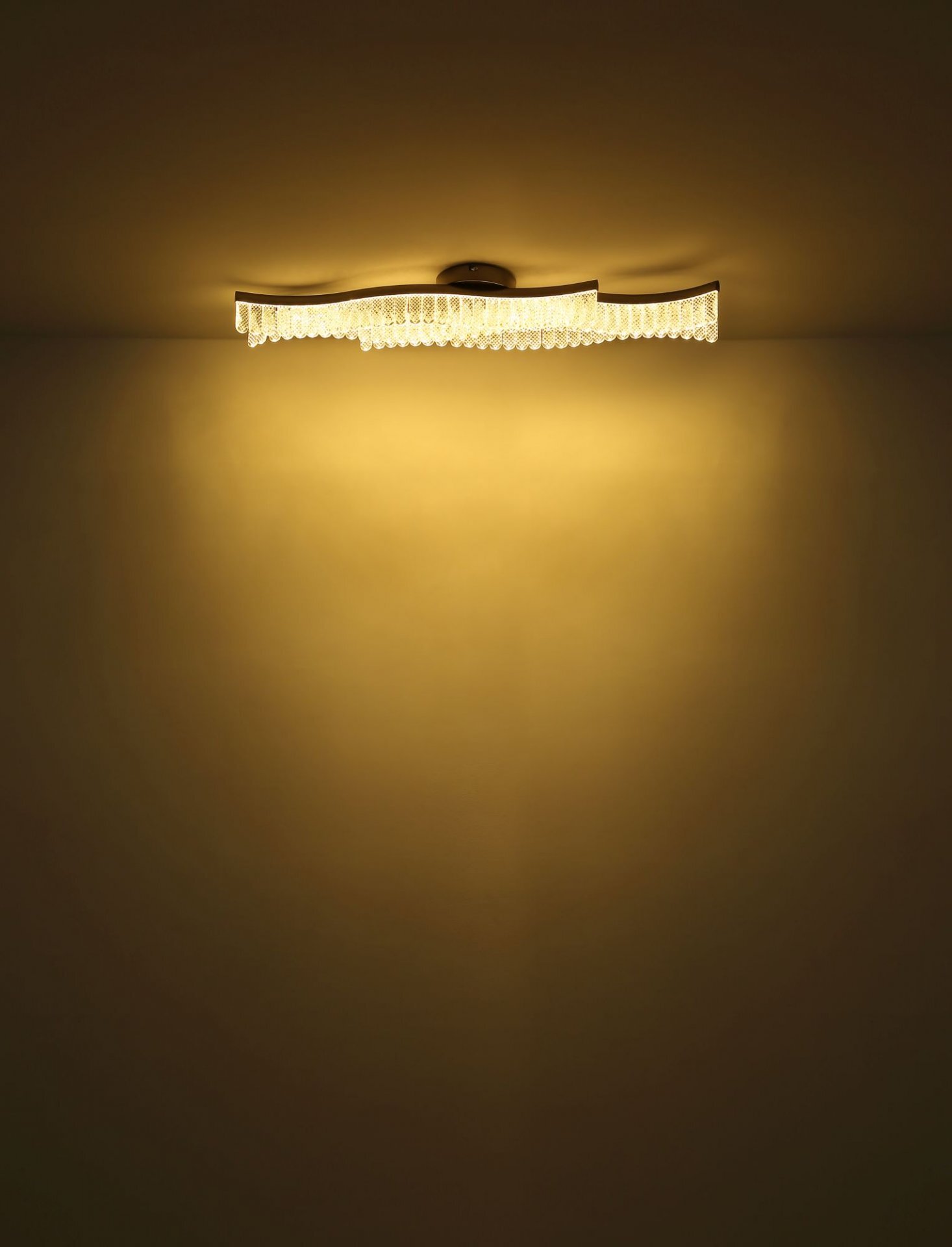 Na obrazie widoczna jest lampa sufitowa emitująca ciepłe, żółte światło, które rozprasza się na suficie i ścianie. Lampa ma falisty kształt i ozdobne elementy przypominające kryształki.