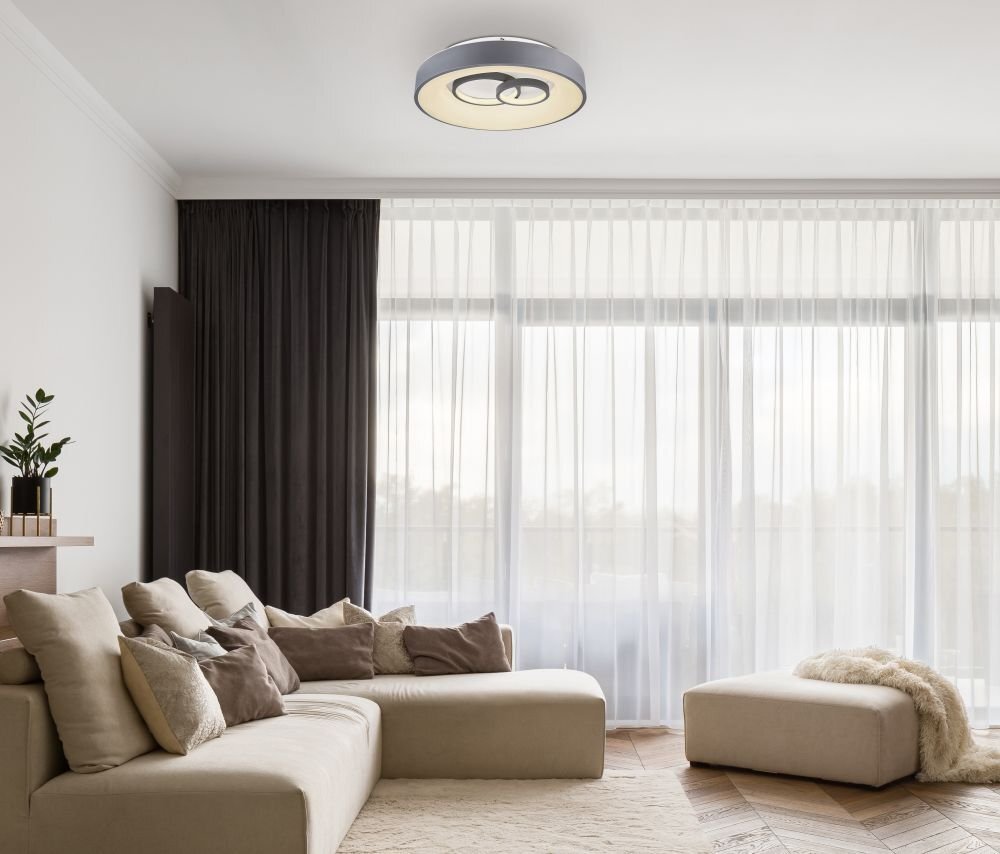 W nowoczesnym salonie znajduje się duża, jasna sofa z poduszkami, a na suficie zamontowany jest okrągły plafon LED. Przez duże okna wpada naturalne światło, a na podłodze leży dywan.