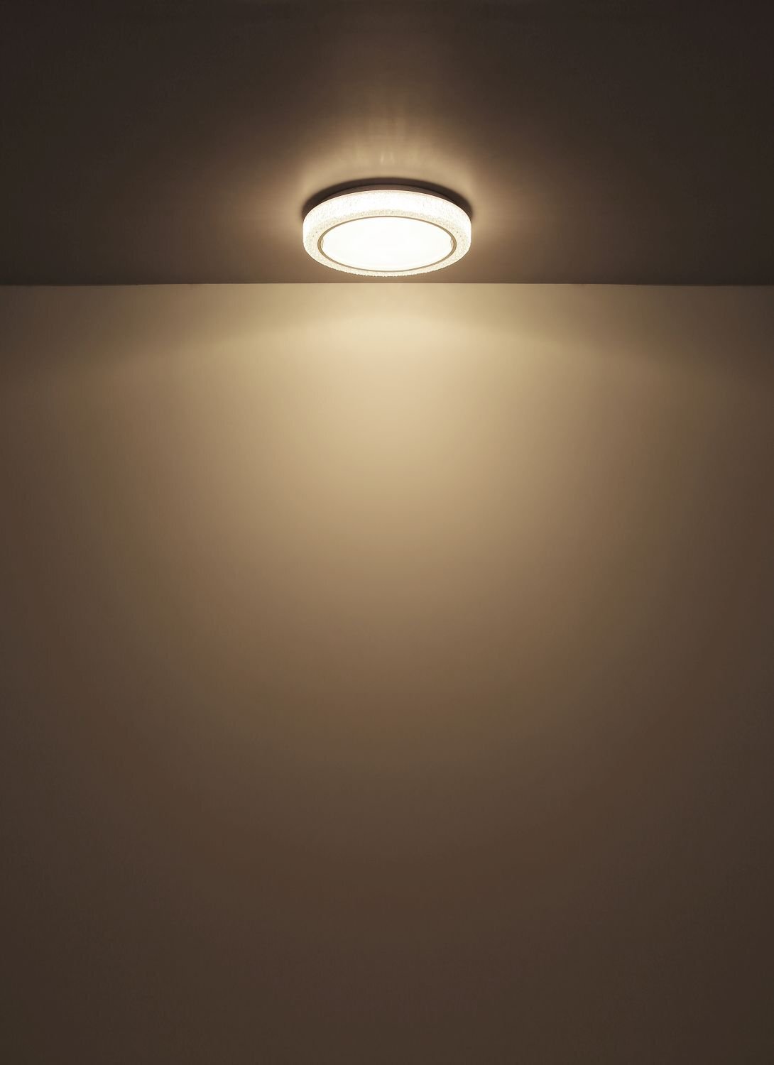 Na suficie zamontowana jest okrągła lampa sufitowa emitująca ciepłe, rozproszone światło. Ściana i sufit są w jasnym, neutralnym kolorze.