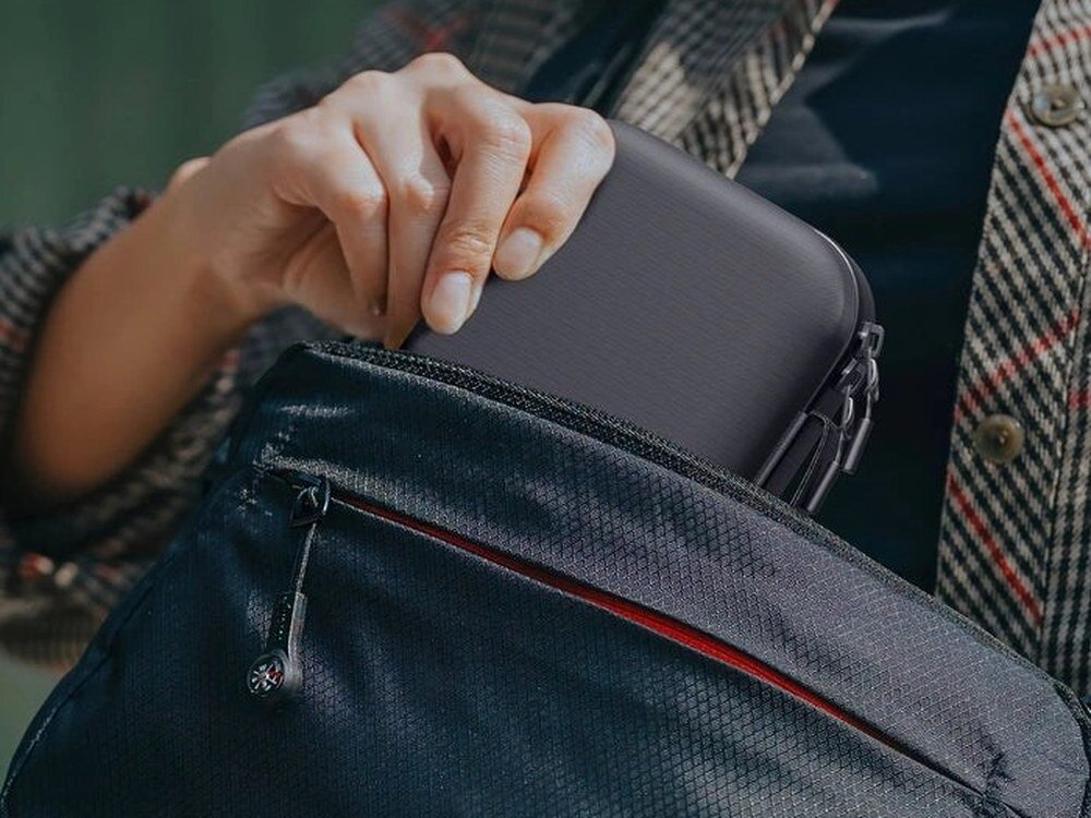 Czarne etui AmazingThing PlayShield wkładane do kieszeni granatowego plecaka, widok z boku, dłoń trzymająca futerał.