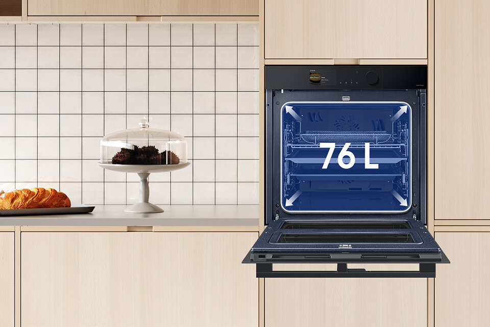 Do kuchni zaaranżowanej w beżowym kolorze wstawiony został piekarnik Samsung. Model NV7B6785KAK, który ma 76 litrów, znajdziesz w sklepach Media Markt. Otwarty piekarnik w zabudowie kuchennej z widoczną pojemnością 76 L na drzwiach. Obok na blacie znajduje się patera z ciastem i talerz z pieczywem.